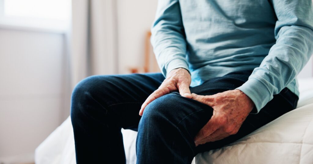 Older man sitting on side of bed holding leg that has muscle cramps