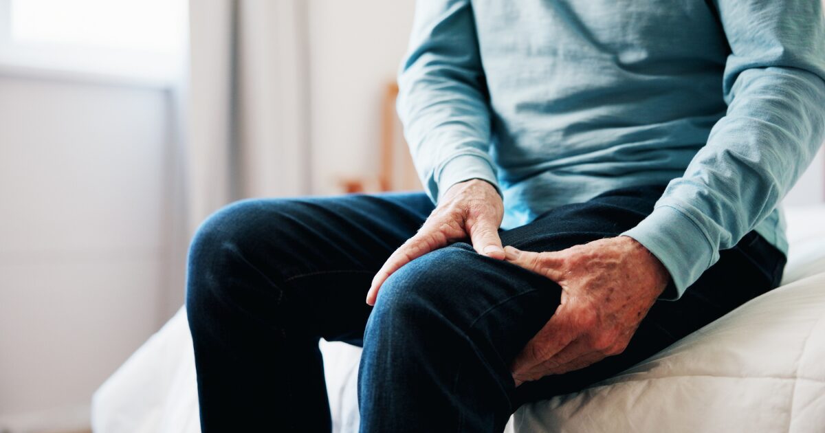 Older man sitting on side of bed holding leg that has muscle cramps