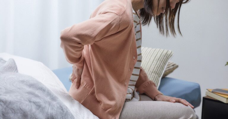 Woman sitting on the edge of her bed holding her lower back in discomfort, illustrating unexpected fibroid symptoms such as back pain and bloating.