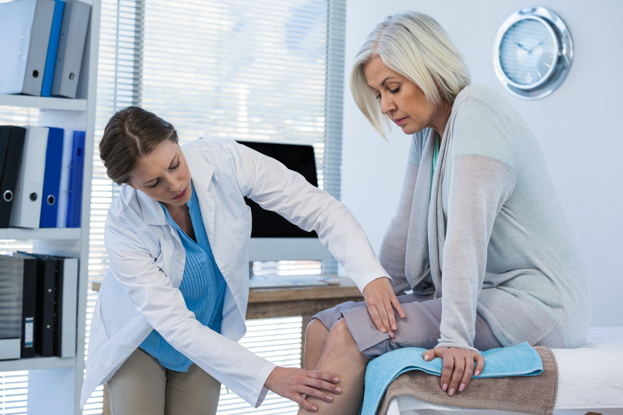 Female healthcare provider examining an older woman’s lower leg during a medical appointment, assessing pain or circulation concerns in a clinical setting.