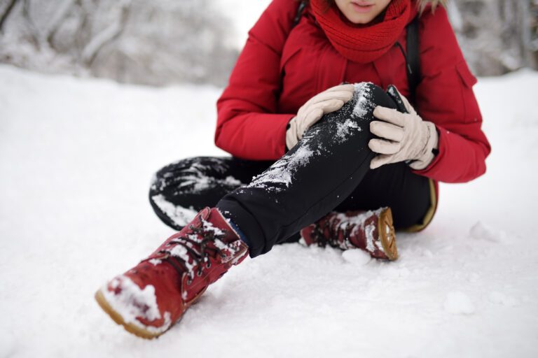 Woman sitting in the snow holding her knee after a fall, wearing winter clothing and red boots with snow on them.