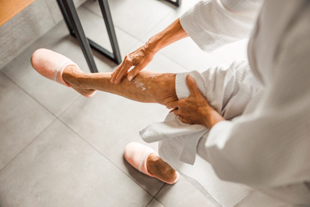 Older woman in a robe applying moisturizer to her lower leg while sitting indoors, with pink slippers visible on the floor.