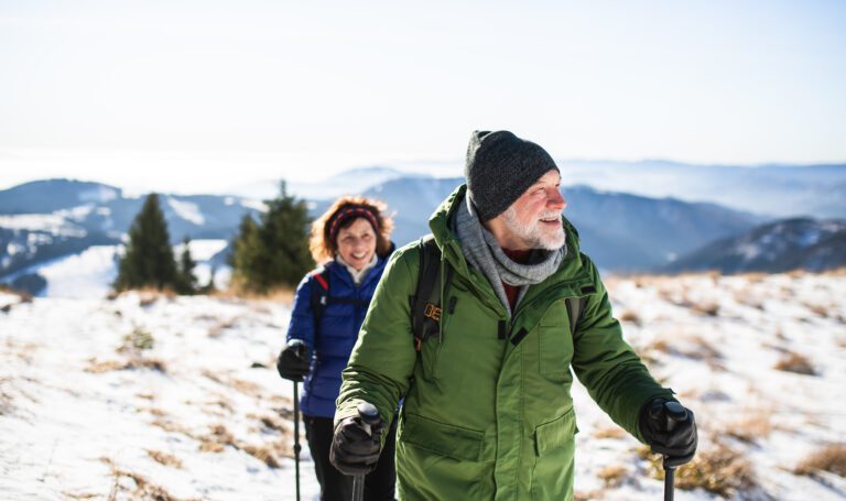 Smiling older couple hiking with trekking poles through a snowy mountain landscape, dressed in warm winter gear.
