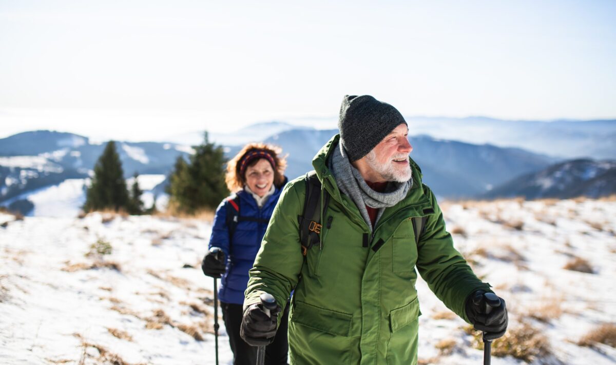 Smiling older couple hiking with trekking poles through a snowy mountain landscape, dressed in warm winter gear.
