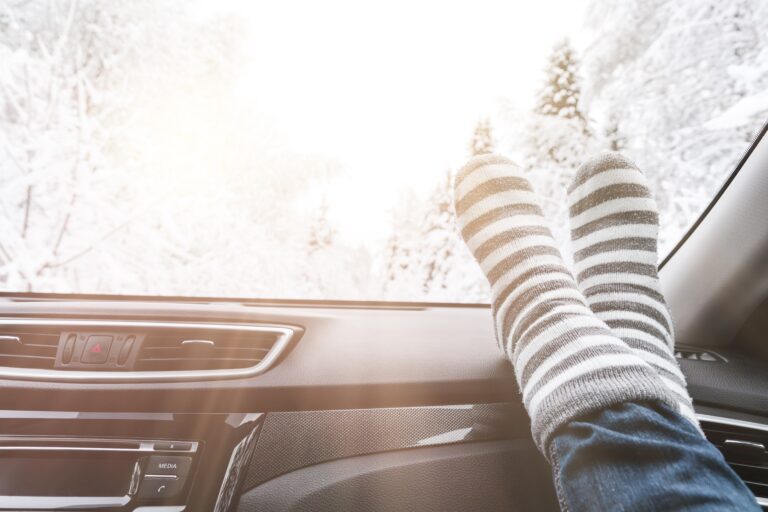 Person wearing striped wool socks with feet propped on a car dashboard, relaxing while looking out at a bright, snowy winter landscape through the windshield.