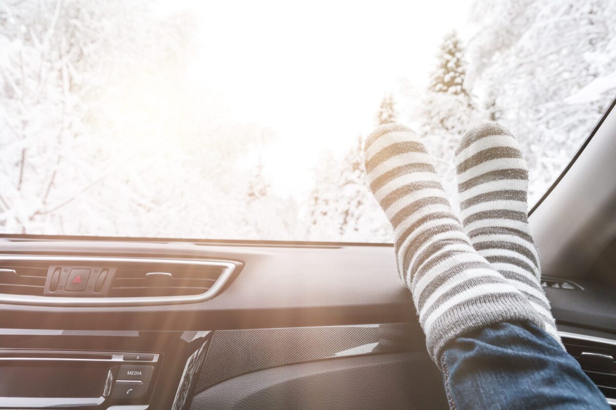 Person wearing striped wool socks with feet propped on a car dashboard, relaxing while looking out at a bright, snowy winter landscape through the windshield.