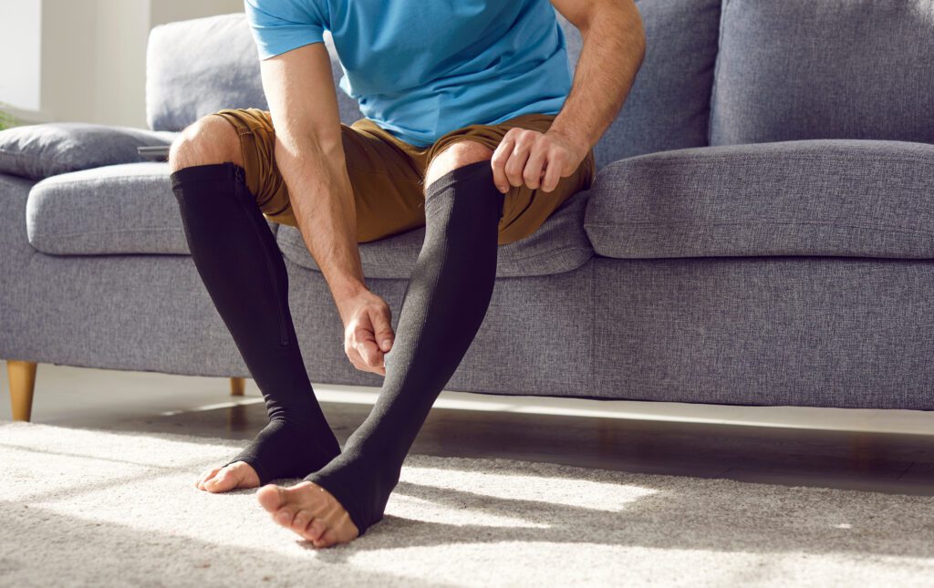 A man sitting on a grey couch adjusting compression stockings on his legs.