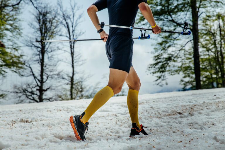 Close-up of a runner’s legs wearing bright compression socks while jogging on a snowy trail, with trekking poles carried behind their back.