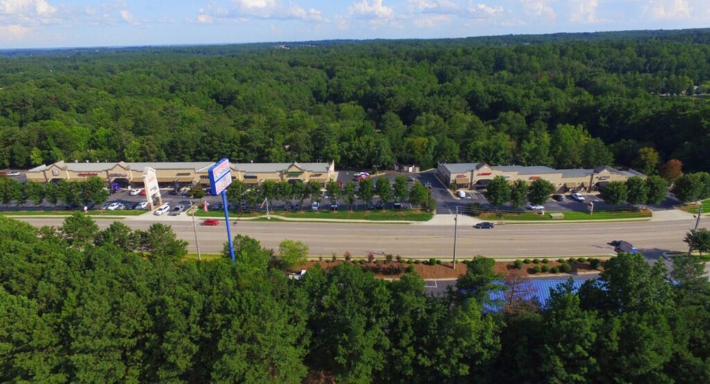 Aerial photo of a commercial plaza along a main road in Douglasville, Georgia, surrounded by dense green trees and parking areas.