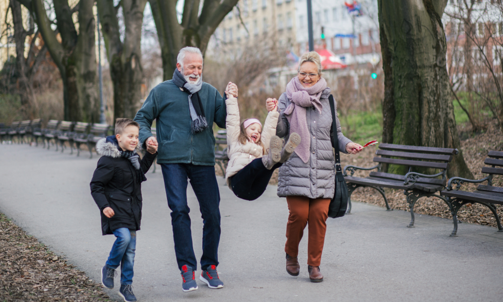 Two adults walking with two children along a park path in cool weather, lifting one child playfully between them while everyone smiles and enjoys the outing.