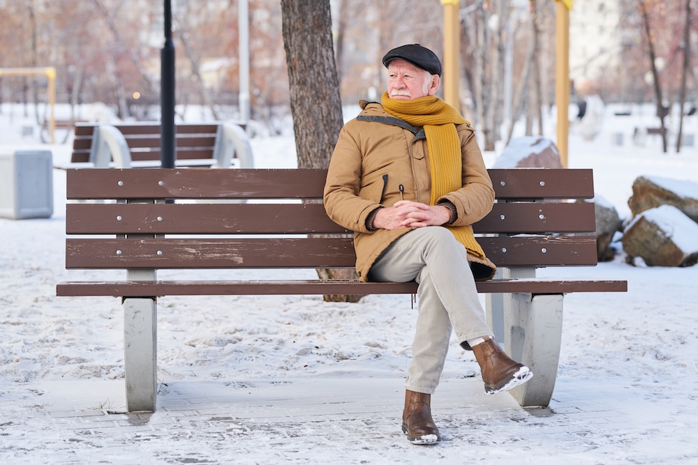 A male senior citizen is sitting on a bench in a snow covered park worried about his aching legs