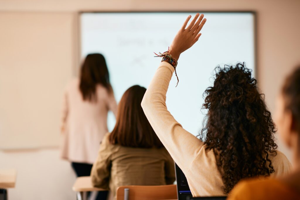 Student with curly hair raising their hand during a classroom lesson, with a teacher writing on a whiteboard at the front of the room.