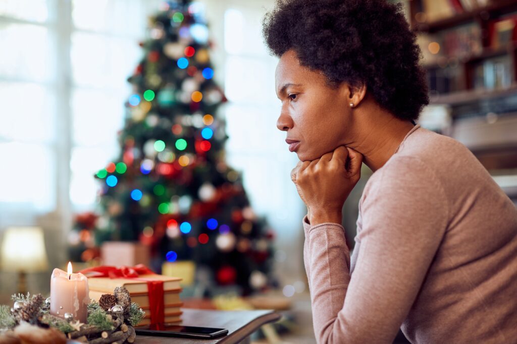 A woman is sitting in with her fist under her chin looking stresses, while a brightly lit Christmas tree is in the background