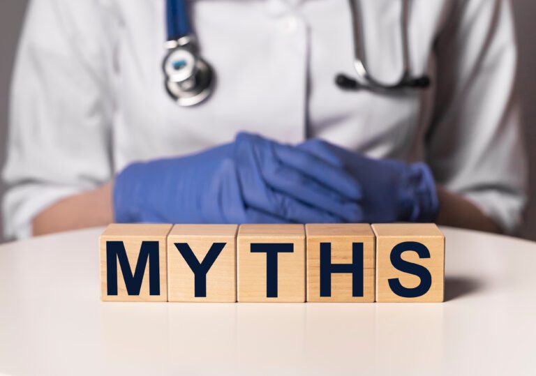 A doctor with gloved hands sitting behind wooden blocks that spell out the word myths