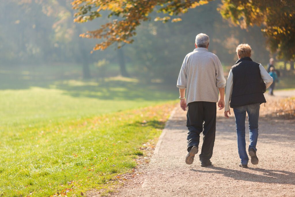 Senior couple walking side by side along a park path on a sunny day, surrounded by green grass and autumn-colored trees.