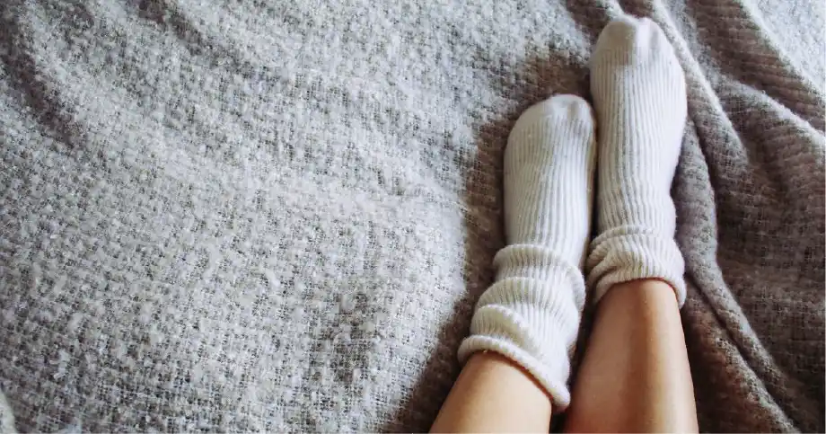 A person's feet with warm socks on and a cozy blanket to protect against cold numb feet