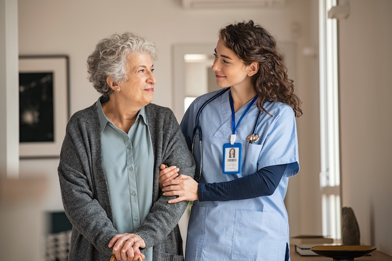 A caring nurse walks a senior woman down a hallway while smiling at each other
