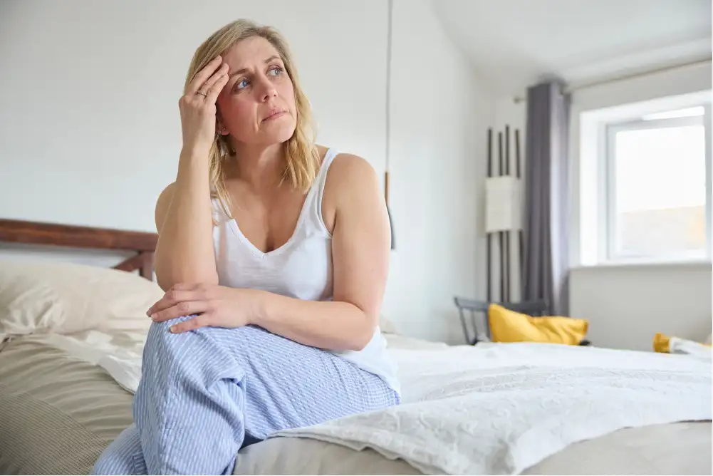 A mid-aged woman sits on the edge of her bed with a concerned look on her face.
