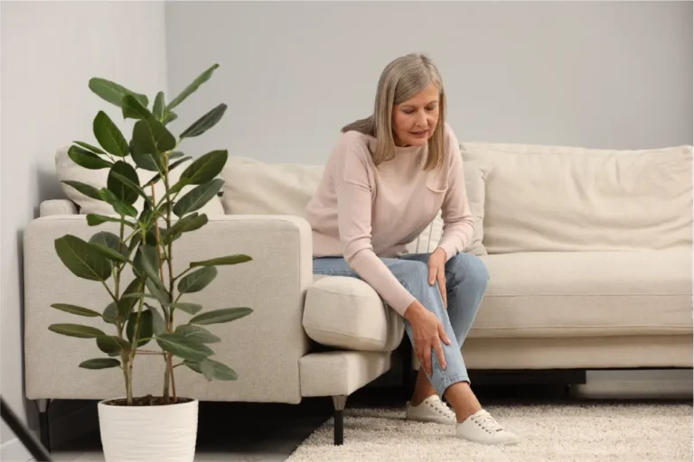 A senior woman sits on a couch bent over holding her lower leg in pain.