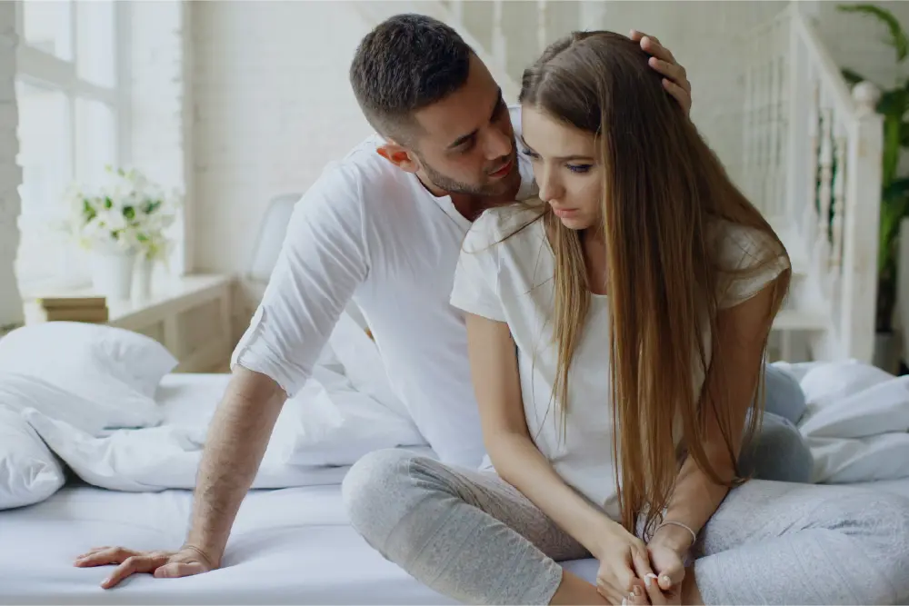 A man and woman are shown sitting on a bed with a sad demeanor
